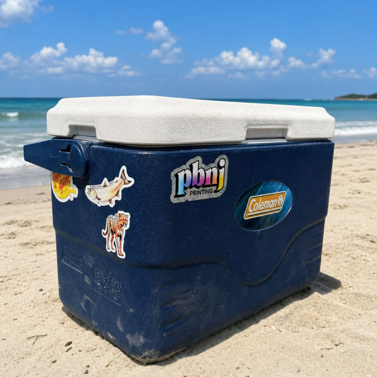 A dark blue cooler with a white lid sits on a sandy beach with the ocean and blue sky in the background. The cooler has several stickers on the side, including a prominent 'Pbnj' logo and a 'Coleman' logo.